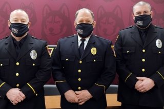 Three university police standing side by side with a red wall behind them.