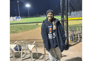 A woman standing outside the fence of a baseball field.