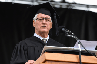 Photo of Dan Cruttenden in graduation garb, speaking at a podium