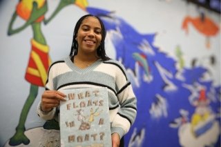 A CU-Lock Haven Early Childhood Education student posing in front of a colorful mural holding the book "What Floats in a Moat?"