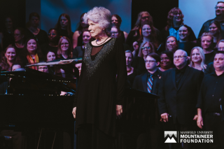 Peggy Dettwiler stands in front of Mansfield alumni choral members with a Mansfield University Mountaineer Foundation logo