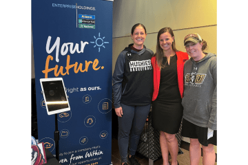 Kelsey Gallagher '12 stands with Husky Basketball coaches at a recent Career Connections Expo in front of a blue Enterprise Holdings roll-up banner