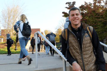 A student leaning against a staircase railing with people walking in the background. 