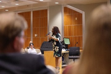 Dr. Carolyn Reid-Brown, wearing a black dress with white flowers, holds a microphone as she speakers with Frederick Douglass scholars