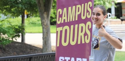 Female student tour guide stands next to Campus Tours signage.