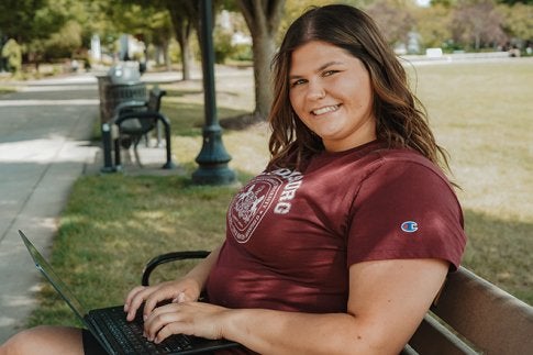 Student employee, Olivia Whitaker, is sitting on a campus bench working on a laptop