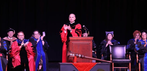 President Hanna stands at a lecturn and applauds the faculty during a convocation ceremony