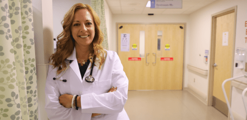 a nurse leaning against a wall, smiling, in front of hospital doors