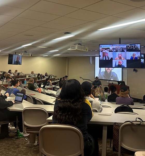 Students sitting in lecture hall with video boards displaying conference panelists.