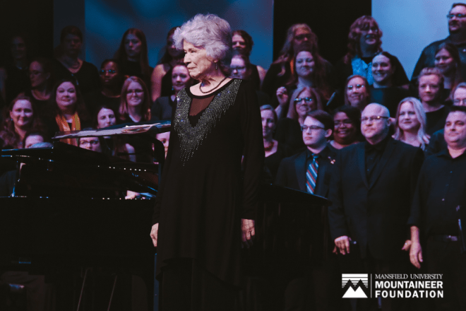 Peggy Dettwiler stands in front of Mansfield alumni choral members with a Mansfield University Mountaineer Foundation logo