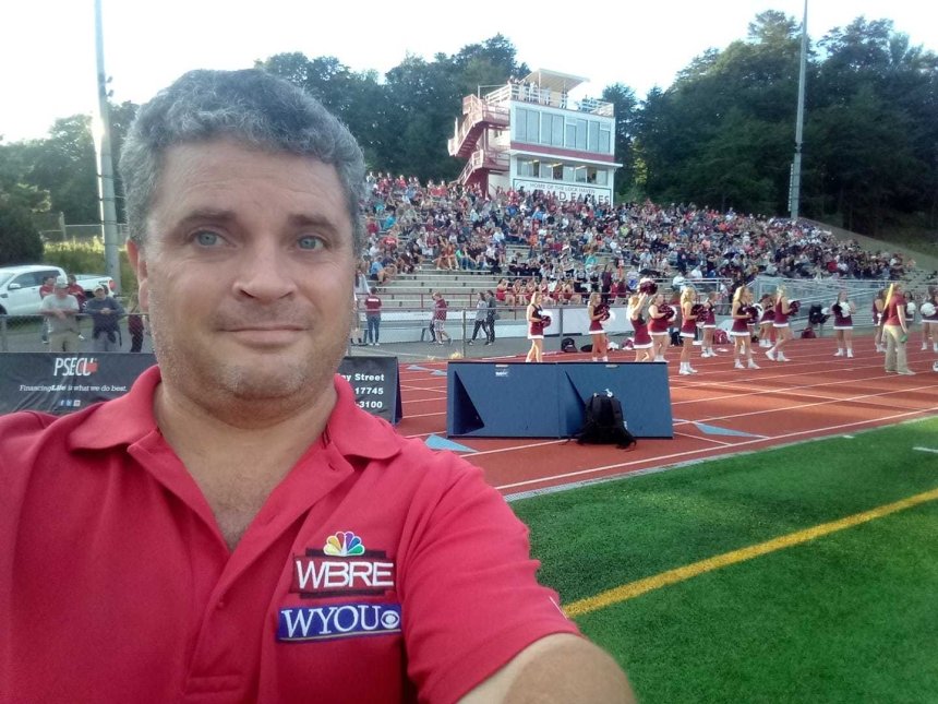 A man taking a selfie in front of bleachers full of people by a field.